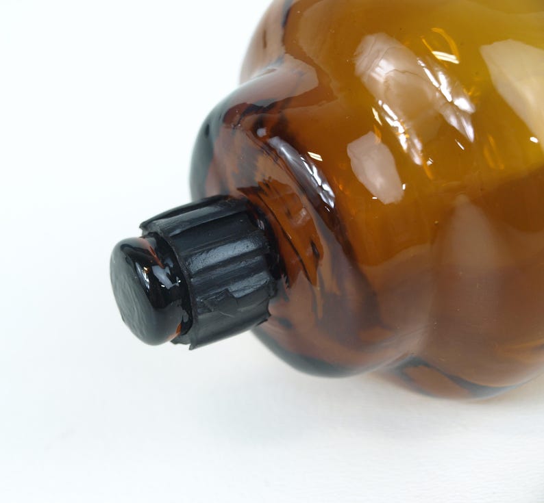 Puede incluir: Una botella de vidrio de color &aacute;mbar transl&uacute;cido con un cuerpo redondeado y bulboso y un cuello corto y estrecho. La botella est&aacute; sellada con una tapa de pl&aacute;stico negra y cil&iacute;ndrica que presenta estr&iacute;as verticales en el lateral para el agarre y una superficie superior lisa y plana.