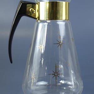 May include: A clear glass pitcher with a gold band around the top and gold starburst designs. It has a black handle and a flared base. A vintage-style beverage container.