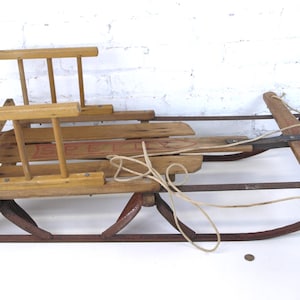 May include: Vintage wooden sled with a metal frame and runners. The sled features a wooden seat and backrest, with a rope for steering. The sled is painted with a red and brown color scheme, and is designed for winter fun.