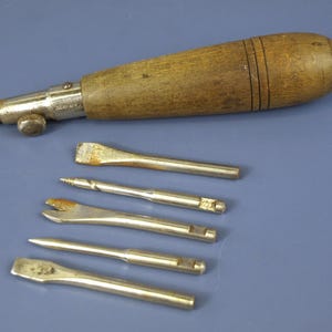 Könnte beinhalten: Ein Vintage-Handwerkzeug mit einem glatten, zylindrischen Holzgriff und einem Metallfutter, begleitet von fünf austauschbaren Metallaufsätzen, darunter ein kleiner Bohrer, eine gabelartige Spitze und drei flache Schraubendreher-Aufsätze.