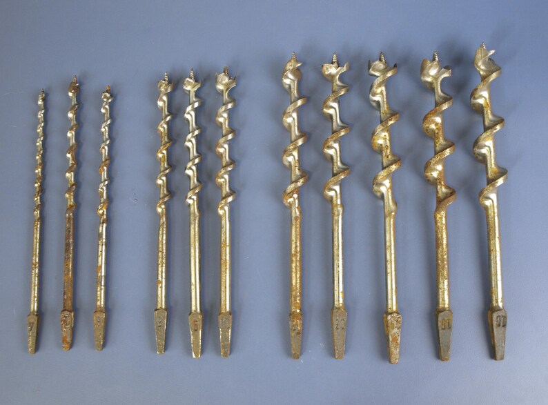 May include: A collection of antique drill bits, varying in size and design. The bits are made of metal with a golden hue and show signs of age and use. The spiral flutes and pointed tips are clearly visible, suggesting their use in woodworking or construction.