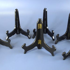 Op de afbeelding: Zes zwarte displaystandaards met een driepotige basis en een hoge, gebogen rug. De standaards hebben een donkere afwerking en zijn ontworpen om borden of andere decoratieve items te bevatten. Sommige standaards hebben gouden accenten.