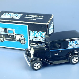 May include: A black 1931 Ford Model A Panel Delivery truck model with white-walled tires and the text "Drag Specialties." The model is next to its box, which features a blue and black design.