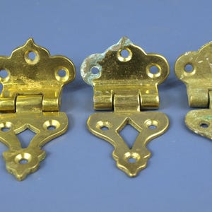 May include: Three vintage brass hinges with decorative cutouts and a penny for size comparison. The hinges are gold-toned with some signs of age and wear, suggesting they are antique or reclaimed hardware.
