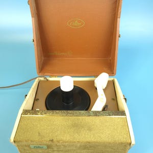 Op de afbeelding: Vintage draagbare platenspeler met een bruine kunstleren behuizing en een goudkleurig luidsprekerrooster. De open deksel onthult een draaitafel met een zwarte plaat en een witte toonarm. De merknaam "Capitol" is zichtbaar.