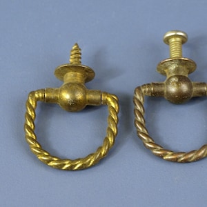 以下が含まれることがあります： ねじれたロープのデザインの2つのアンティーク真鍮引き出しプル。1つはネジ、もう1つはボルトが付いています。プルは金色で、青灰色の背景にあります。これらのプルは、おそらく家具の修復用です。