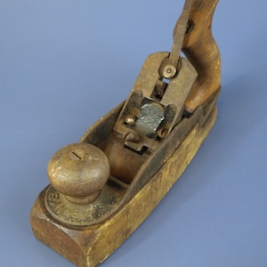 May include: A vintage Bailey hand plane featuring a weathered wooden base, a rounded wooden front knob, and a contoured wooden rear handle. The metal mechanism includes a central blade assembly with visible screws and a lever adjustment component.