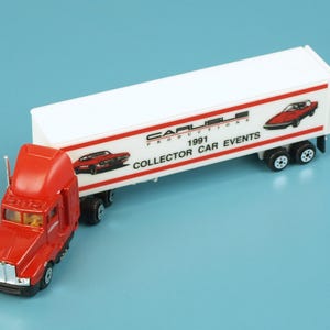 May include: A red toy semi-truck with a white trailer. The trailer has red stripes and images of classic cars. Text on the trailer reads "CARLISLE PRODUCTIONS 1991 COLLECTOR CAR EVENTS."