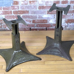 May include: Two industrial metal table bases with a dark olive green finish and a four-point flared base. Each base features a vertical support column with an adjustable top plate and the word WELFARE embossed on one side of the base structure.