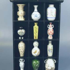 Puede incluir: Una estantería negra exhibe doce pequeños jarrones decorativos. Los jarrones, de diversas formas, presentan detallados diseños florales y geométricos en colores como blanco, azul, verde y dorado. El conjunto se encuentra sobre un fondo azul claro.
