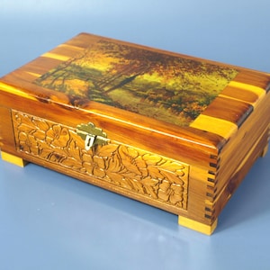 May include: A rectangular wooden box with a hinged lid. The lid features a landscape painting. The front of the box has a carved floral design. The box is made of light and dark brown wood with a brass clasp.