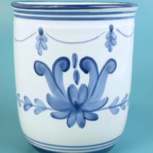 May include: White ceramic vase with blue floral design. The vase features a blue floral motif in the center, with a garland design and blue stripes at the top and bottom. The background is a solid light blue.