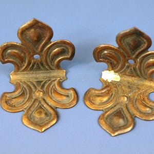 May include: Two antique bronze-colored metal decorative fleur-de-lis shaped hardware pieces. Each piece has a central hole and ornate, symmetrical scrollwork design. The hardware has a distressed finish, suggesting age and use.