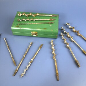 May include: A collection of vintage auger drill bits and a green metal case. The drill bits are various sizes and have a spiral design. The case is rectangular with a clasp. The tools are arranged on a blue surface.