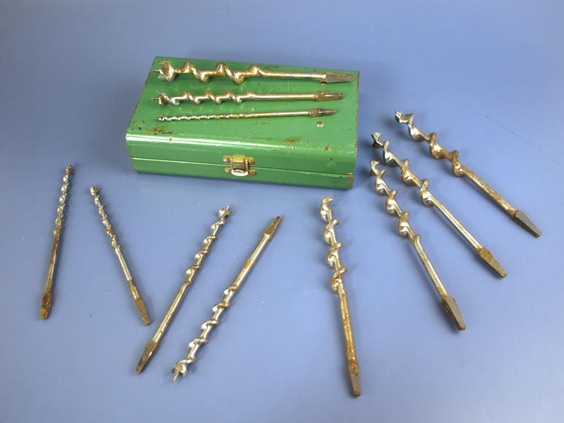 May include: A collection of vintage auger drill bits and a green metal case. The drill bits are various sizes and have a spiral design. The case is rectangular with a clasp. The tools are arranged on a blue surface.