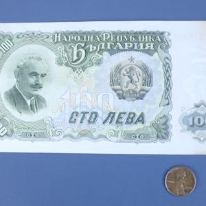 May include: A Bulgarian 100 Leva banknote with a portrait of a man, text in Cyrillic, and a coat of arms. A penny is next to the banknote for size comparison. The banknote is on a blue surface.