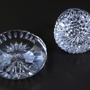 May include: Two clear, faceted crystal decorative objects. One is a shallow bowl shape, and the other is a rounded, dome shape. Both have intricate starburst patterns cut into the crystal, reflecting light. The objects are on a dark background.