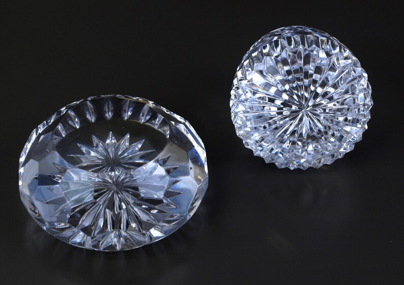 May include: Two clear, faceted crystal decorative objects. One is a shallow bowl shape, and the other is a rounded, dome shape. Both have intricate starburst patterns cut into the crystal, reflecting light. The objects are on a dark background.