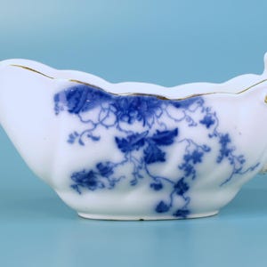 May include: White porcelain gravy boat with a blue floral design. The boat has a scalloped rim with gold trim and a handle. The blue floral pattern is a vine-like design. The gravy boat is on a light blue background.