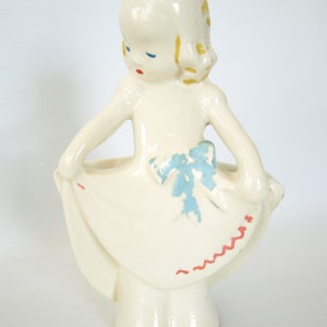 May include: A white ceramic figurine of a girl with blue eyes and yellow hair wearing a white dress with a blue bow and red stitching. The girl is holding her dress up and has a hole in the back for planting.