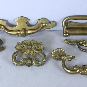 以下が含まれることがあります： 装飾的なデザインが施されたアンティークな真鍮製の引き出しノブが6つあります。ノブは、長方形のノブ、湾曲したノブ、花柄のノブなど、さまざまな形とサイズがあります。