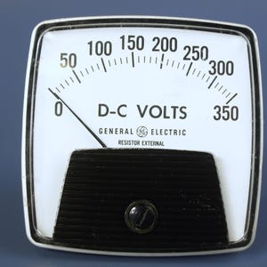 May include: A square General Electric D-C Volts analog meter with a white face, black markings, and a needle pointing to zero. The gauge features a black ribbed lower section with a central screw and numerical increments ranging from 0 to 350.