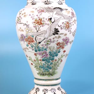 May include: A white ceramic vase with a flared base and a rounded top. The vase is decorated with hand-painted cranes, flowers, and foliage in black, red, pink, green, and blue. The rim and base have decorative patterns. A decorative item.