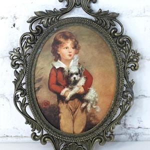 Puede incluir: Un marco de fotos ovalado de latón con detalles ornamentados. El marco contiene una impresión vintage de un niño pequeño sosteniendo un perro blanco pequeño.