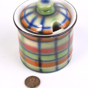 Może przedstawiać: Ceramiczna cukiernica z pokrywką i otworem na łyżeczkę. Miska ma wzór w kratę w kolorze zielonym, pomarańczowym i czerwonym. Pokrywka ma pasujący wzór w kratę z zielonym guzikiem. Obok miski leży moneta.