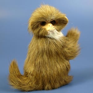 May include: A plush monkey figurine with brown fur, a light-coloured face, and dark eyes. The monkey has a tuft of white fur under its chin and is posed with one arm raised.