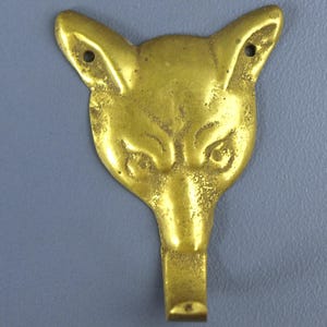 May include: A gold-colored metal wall hook shaped like a fox head. The fox head features pointed ears with a small circular hole in each ear for mounting purposes. The hook extends downward from the chin of the fox, providing a functional hanger.