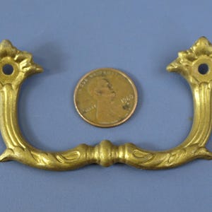 以下が含まれることがあります： 金色の装飾的な引き出しの取っ手。 花柄のデザインで、U字型で装飾的なディテールと2つの取り付け穴があります。 サイズの参考としてペニーが含まれています。 家具の修復やヴィンテージプロジェクトに適した金具です。