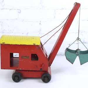 May include: Vintage red and yellow toy crane with a teal bucket. The crane has a red body with a yellow roof and black wheels. The bucket is suspended by a wire. The word "Structo" is visible on the side of the crane.