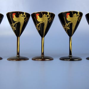 Puede incluir: Cinco copas de cóctel negras con tallos y bordes dorados. Cada copa presenta un diseño de gallo dorado con una cresta roja. Las copas están dispuestas en fila sobre una superficie azul claro.