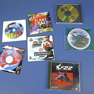 May include: An assortment of computer game CDs, including "NASCAR HEAT," "PLANE CRAZY," "NASCAR THUNDER 2004," and "F22." The CDs are in their original cases, with colorful graphics and game titles visible. The background is a solid blue surface.