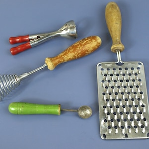Può includere: Un set di quattro utensili da cucina composto da uno scavino in metallo con manici rossi, una frusta in filo metallico con manico in legno usurato, un piccolo scavino con manico verde e una grattugia rettangolare in acciaio inox con manico.