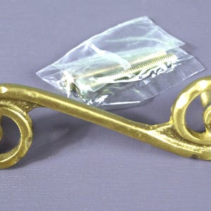 May include: A gold-colored, decorative drawer pull with a scroll design. The hardware has a textured surface and is accompanied by screws in a clear plastic bag. The drawer pull is a decorative element for furniture.