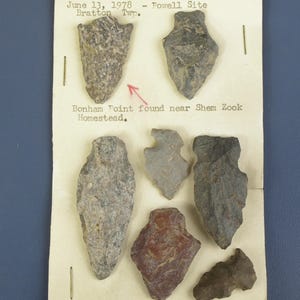 May include: A collection of seven antique arrowheads of varying sizes and shapes, displayed on a cream-colored paper. The arrowheads are primarily gray, brown, and reddish-brown. Text on the paper indicates the date and location of the find.