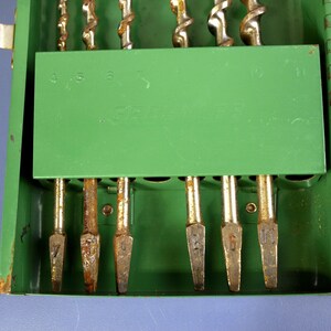 May include: A green metal drill bit case with a selection of drill bits. The case has a hinged lid and a green interior with slots to hold the drill bits. The drill bits are various sizes and have a silver and gold color.