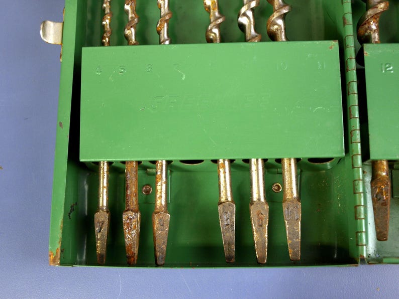 May include: A green metal drill bit case with a selection of drill bits. The case has a hinged lid and a green interior with slots to hold the drill bits. The drill bits are various sizes and have a silver and gold color.