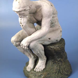 May include: A white statue of a seated figure, with the head resting on the hand, in a contemplative pose. The statue is on a green and brown textured base. The figure is painted in a weathered style, with visible details.