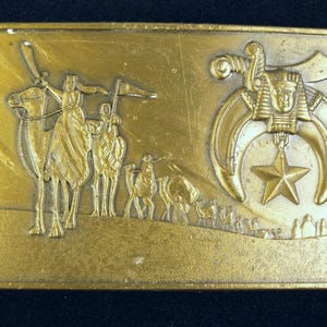 May include: A rectangular gold-colored belt buckle with embossed imagery. The left side depicts figures riding camels, while the right side features a crescent moon, star, and pharaoh's head. The buckle has a vintage aesthetic.