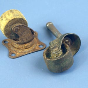 May include: Two antique furniture casters. One caster is mounted on a rusty, star-shaped plate. The other caster has a metal stem. Both casters have a worn, aged appearance with a yellowed wheel.