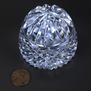 May include: A clear, cut-crystal trinket box with a domed lid. The box features a diamond-cut pattern on the lower portion and a starburst design on the lid. A penny is next to the box for size comparison.