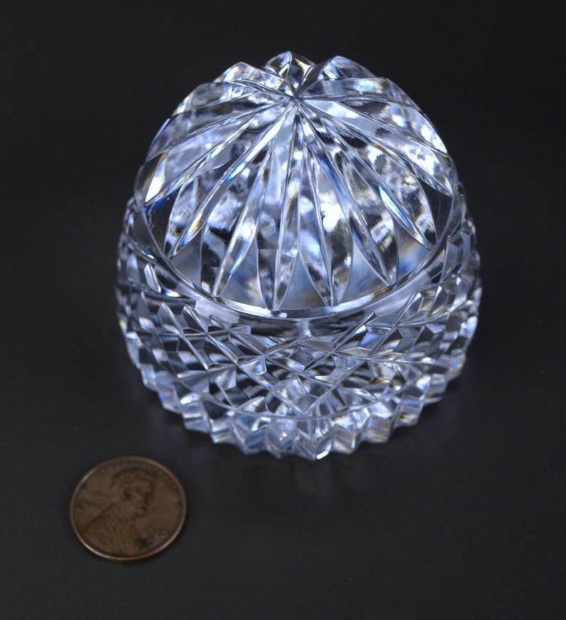 May include: A clear, cut-crystal trinket box with a domed lid. The box features a diamond-cut pattern on the lower portion and a starburst design on the lid. A penny is next to the box for size comparison.