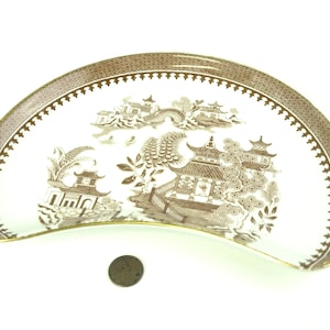 以下が含まれることがあります： 曲線的な形状の茶色と白の磁器皿。お皿には、塔や木々が描かれた詳細な風景画が描かれています。縁には装飾的な茶色の模様があります。お皿の隣にはコインがあります。