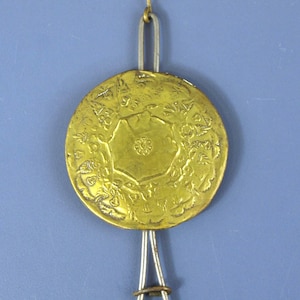 Puede incluir: Un péndulo de reloj circular de metal dorado con un patrón floral en relieve y un motivo de flor central. El disco está suspendido de una varilla de metal plateado con un pequeño gancho en la parte superior y un anillo deslizante abajo.