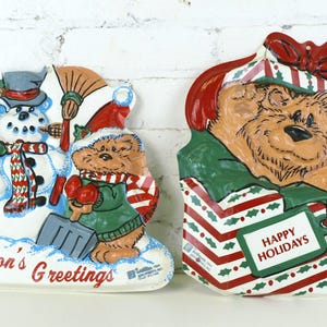 May include: Two ceramic Christmas decorations. One features a snowman and two bears with the text "Season's Greetings". The other shows a bear in a gift box with the text "Happy Holidays". Both are painted in bright colors.