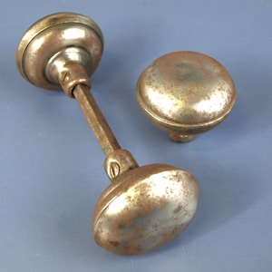 May include: A set of two round, metallic door knobs with a weathered, tarnished finish. One knob is attached to a long, square-shaped metal spindle, while the other knob is separate. Both knobs feature visible screws on the neck.