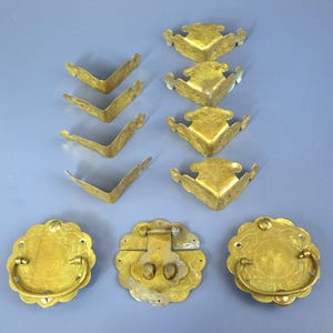 May include: A collection of brass hardware including four L-shaped corner protectors with decorative edges, four ornate corner brackets with engraved patterns, two circular drawer pulls with swinging handles, and one central latch mechanism.
